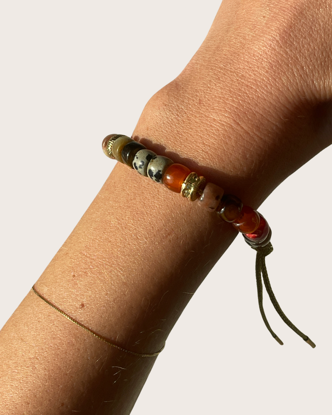 A close-up of a person's wrist wearing a beaded gemstone bracelet with earthy tones, including orange, brown, black, and white marbled beads, accented by gold-toned spacers and a dark cord tassel, against a plain light background.