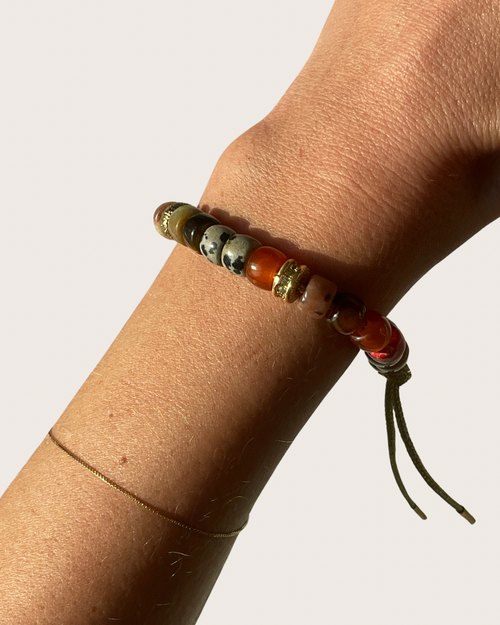 A close-up of a person's wrist wearing a beaded gemstone bracelet with earthy tones, including orange, brown, black, and white marbled beads, accented by gold-toned spacers and a dark cord tassel, against a plain light background.